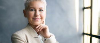 Elegant middle aged businesswoman with stylish pixie hair standing by window in office, touching face, thinking about business issues, looking at camera with confident smile. Success, career and job