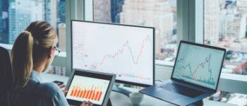 Back view of business woman sitting at panoramic skyscraper office desktop front PC computer with financial graphs and statistics on monitor.Analysis of digital market and investment in block chain
