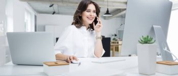 Young businesswoman sitting at her desk talking on phone and working on computer. Female executive working in modern workplace.
